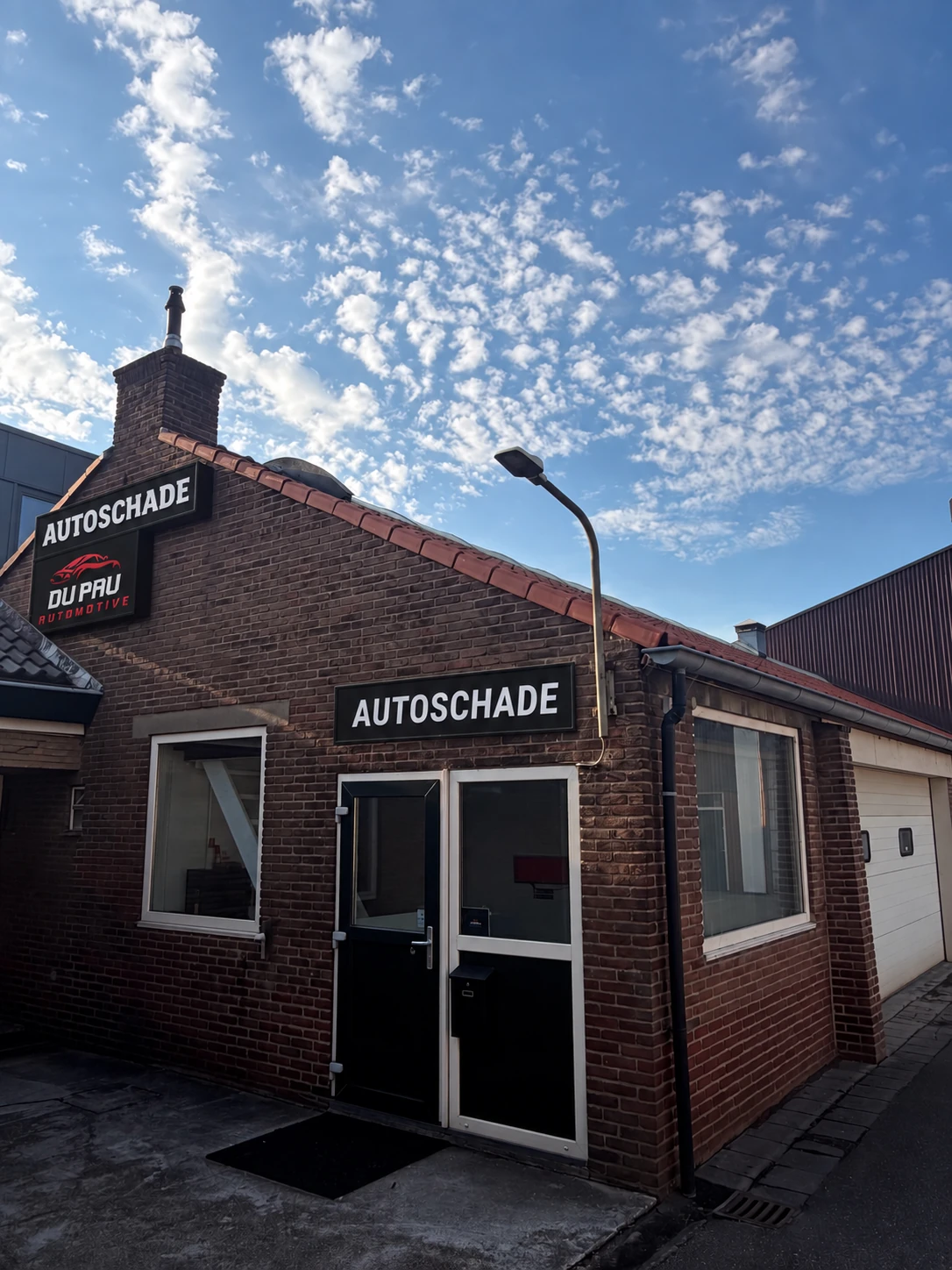 Het pand van DU PAU AUTOMOTIVE onder een blauwe hemel, met het logo, de ingang en de borden Autoschade
